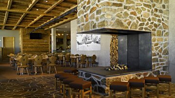 Ein modernes Restaurant mit Holzdekor und einer markanten Steinmauer. In der Mitte steht ein offener Kamin, umgeben von gemütlichen Sitzplätzen.